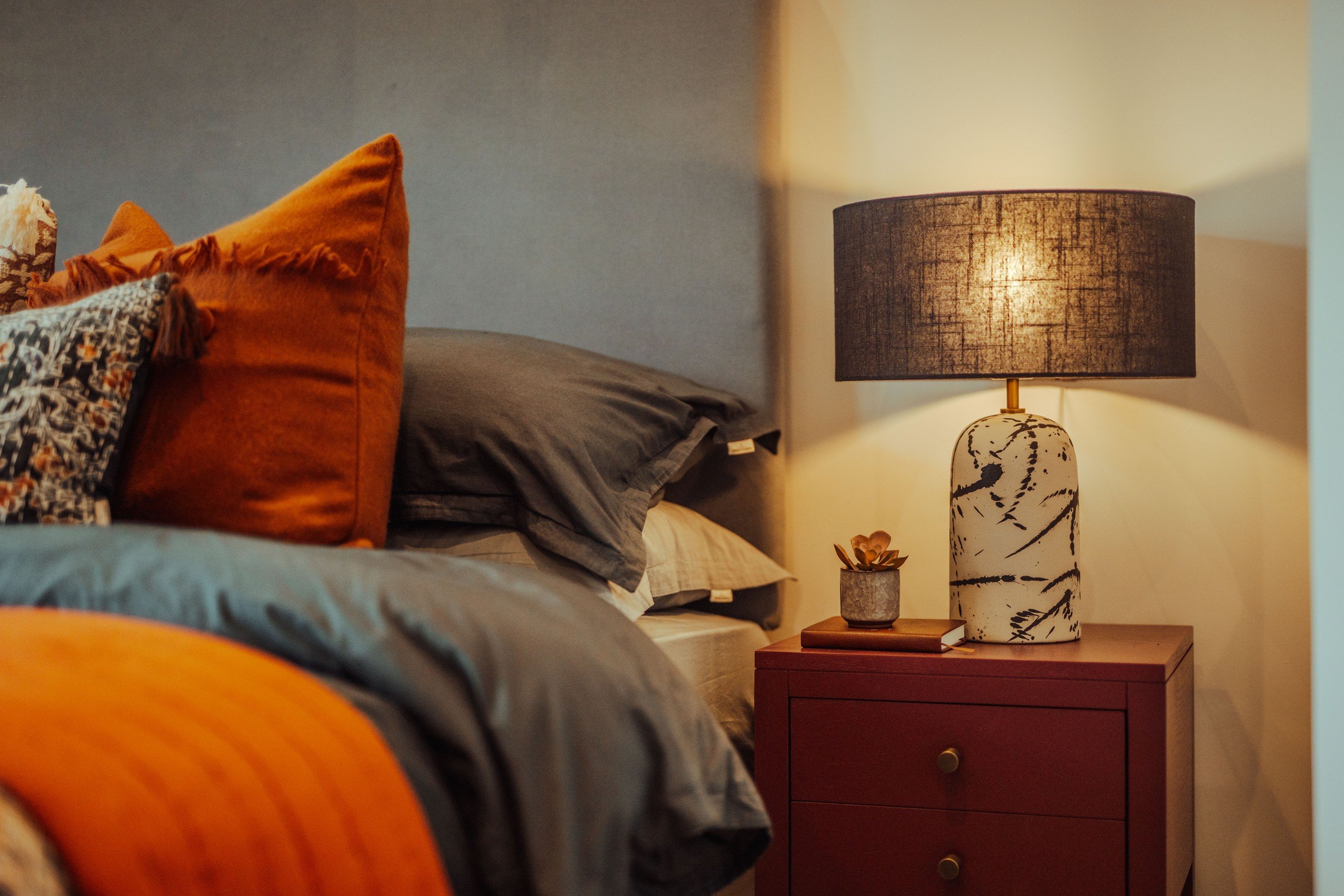 Cozy bedroom with a lit lamp on a red nightstand, gray sheets, and vibrant orange pillows. Soft, warm lighting and a peaceful mood.