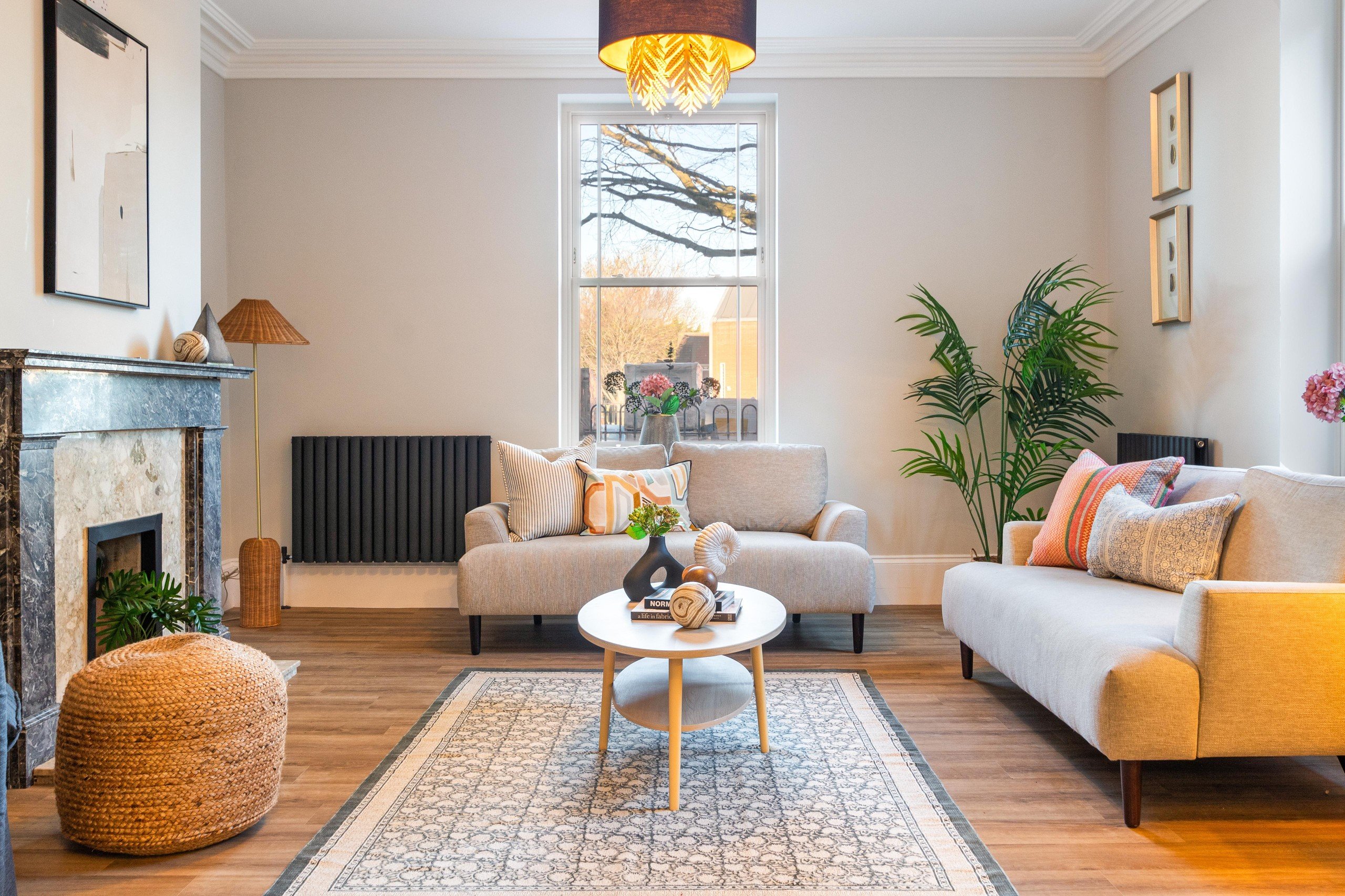 Modern living room with beige sofas, colorful pillows, a round table with books, and a patterned rug. A plant and fireplace are in the background.