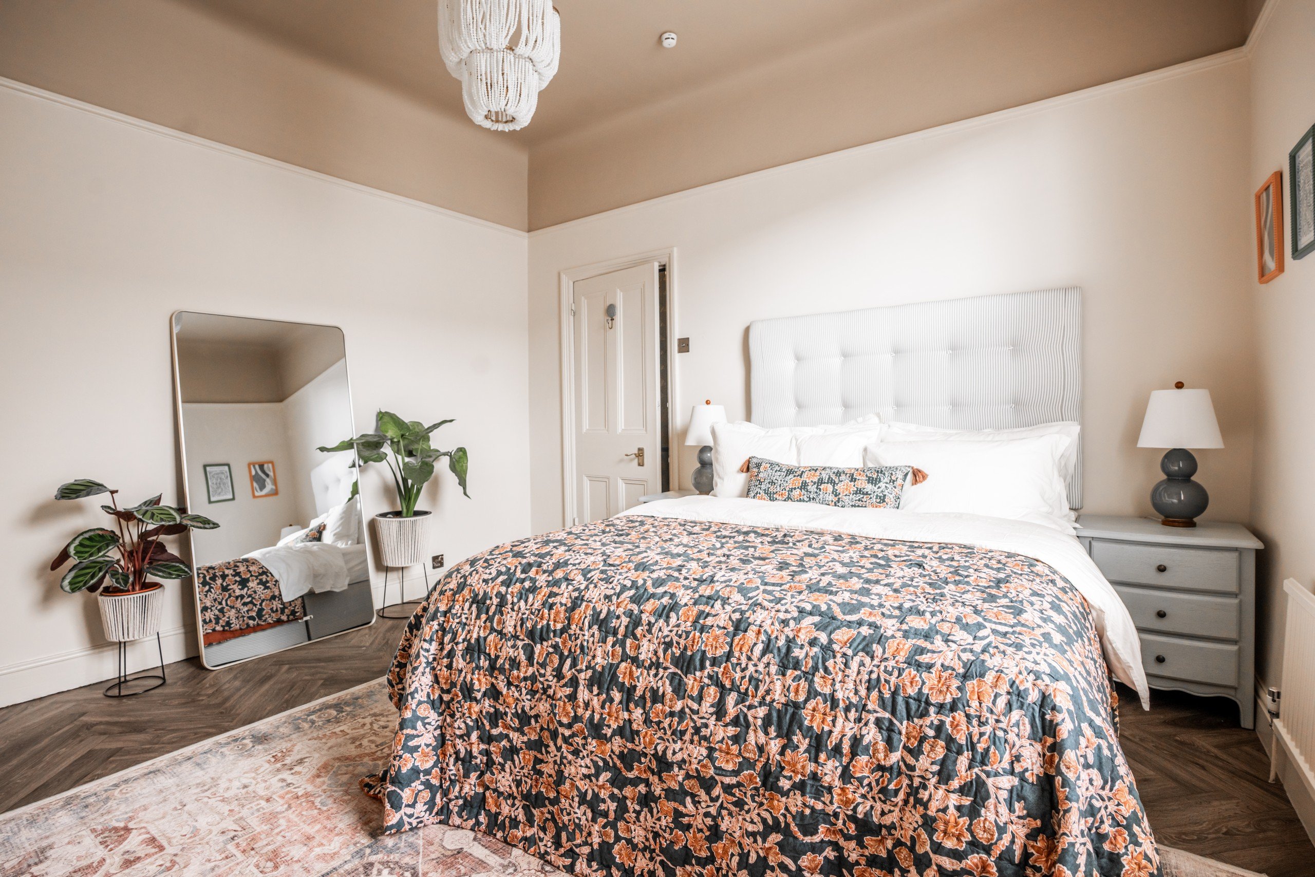 Cozy bedroom with a floral bedspread, white headboard, and plants by a mirror. Soft lighting and a neutral-toned rug enhance a calming ambiance.