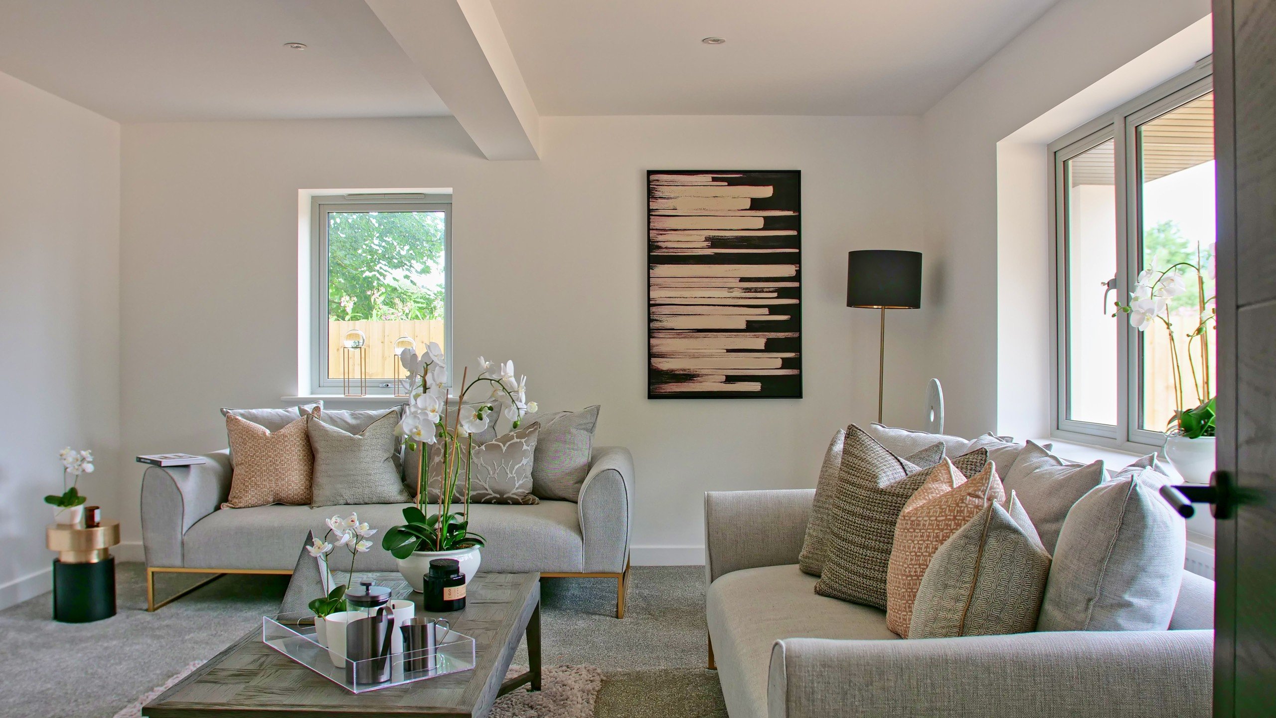 Modern living room with grey sofas, beige cushions, and orchids. Abstract art on white walls, large windows, a black lamp, and a coffee table.