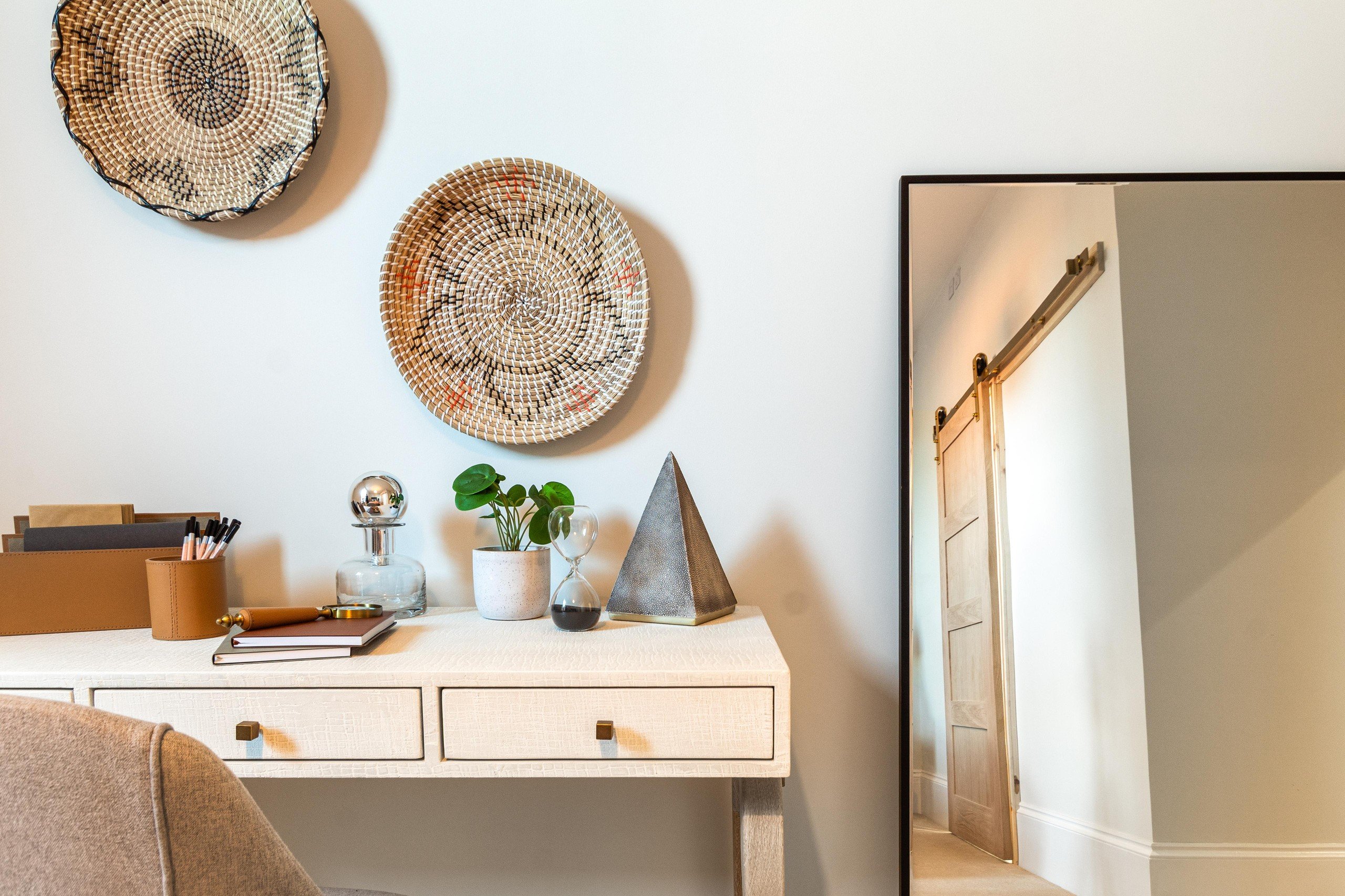 A tidy desk with decor: notebooks, pens, a plant, and an hourglass. Woven baskets hang above; a mirror reflects a wooden door. Neutral tones.